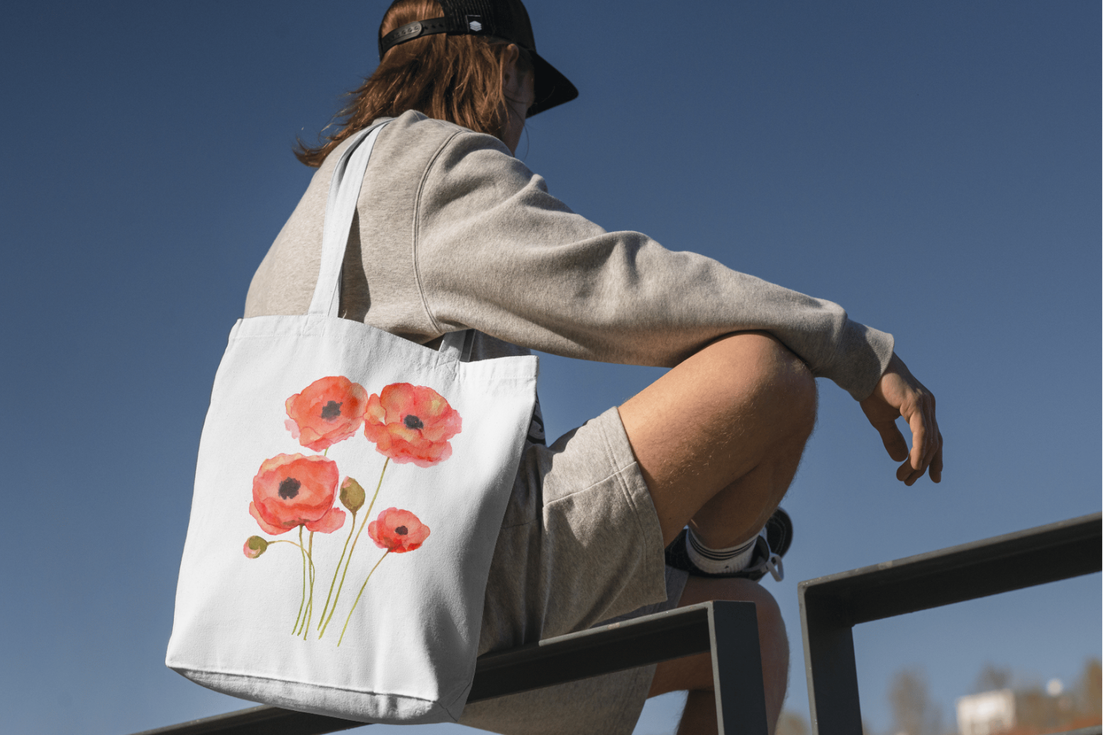 Person sitting outdoors with a tote bag featuring red flowers.